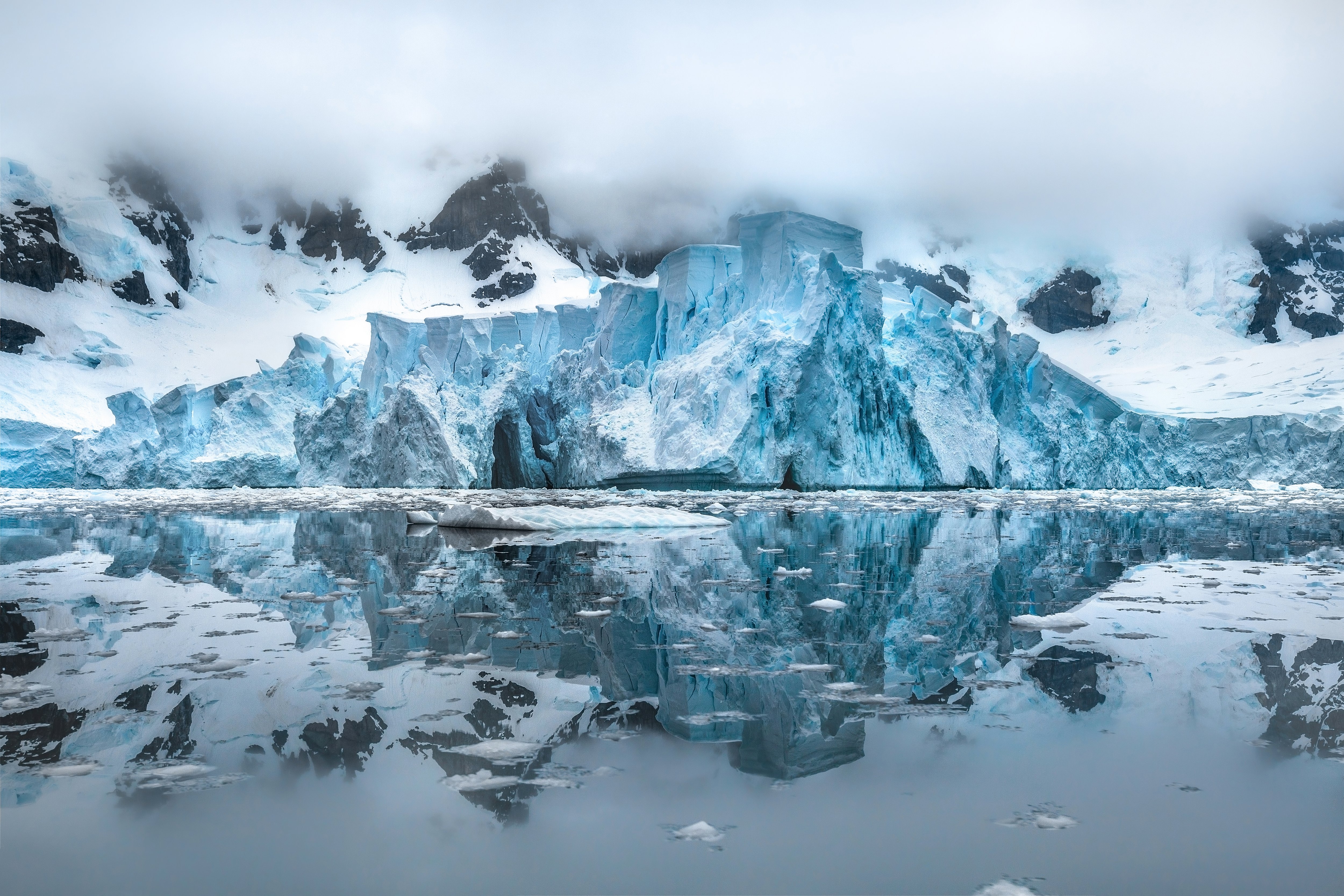 A wide landscape photograph of the glowing blue Suárez Glacier at Paradise Harbour on the Antarctic Peninsula. The massive ice formation is flanked by snow-covered dark mountains under a heavy blanket of low-hanging white fog and clouds. The glassy, mirror-like dark water in the foreground reflects the intricate patterns of the glacier and floating fragments of ice.