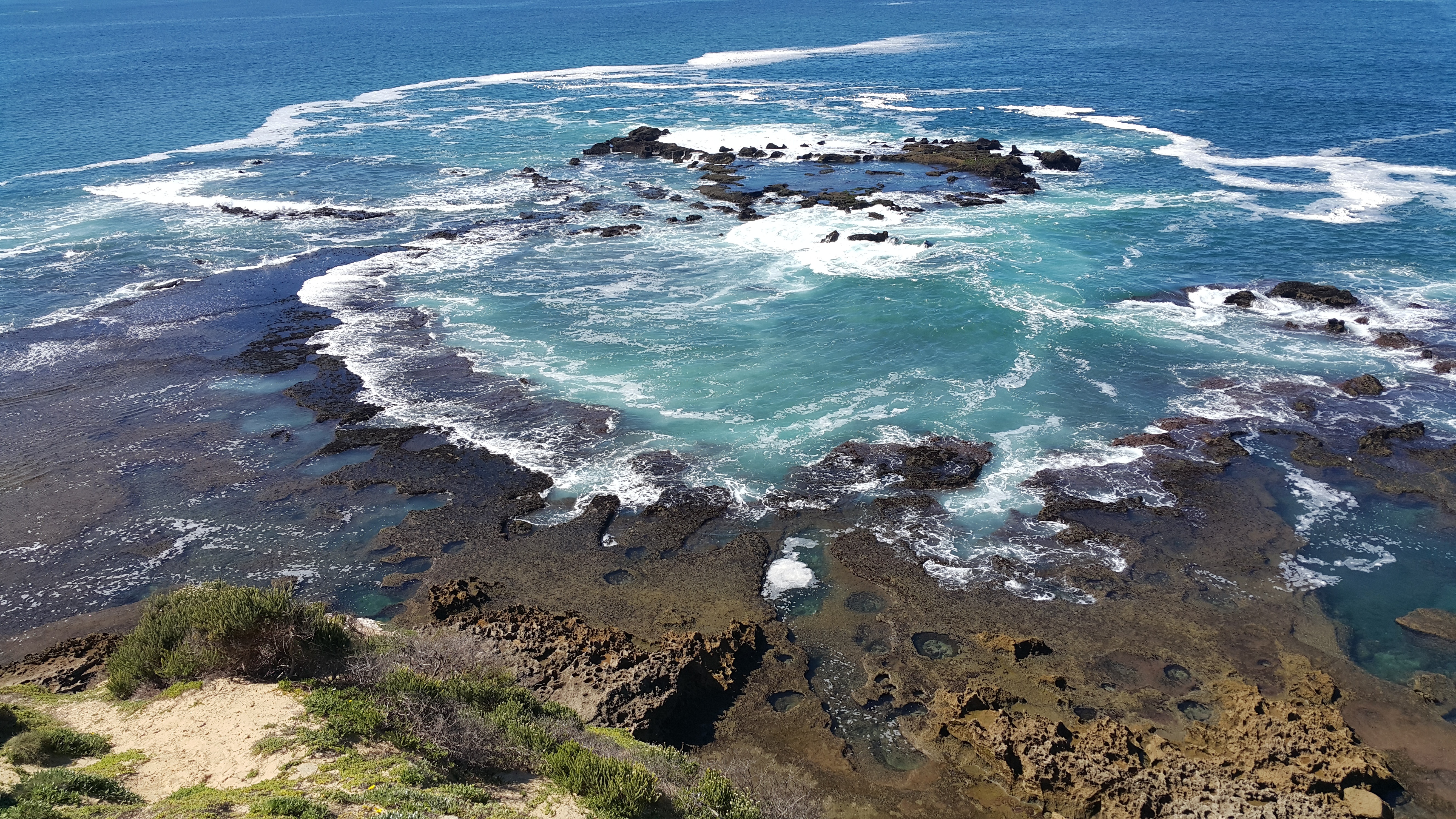 A high-angle, scenic view looking down from a grassy cliffside onto a large, natural rocky tide pool formation in the ocean. The deep blue sea flows into the central pool, creating swirling turquoise water and rings of bright white seafoam as waves crash gently against the dark, textured rock reef.