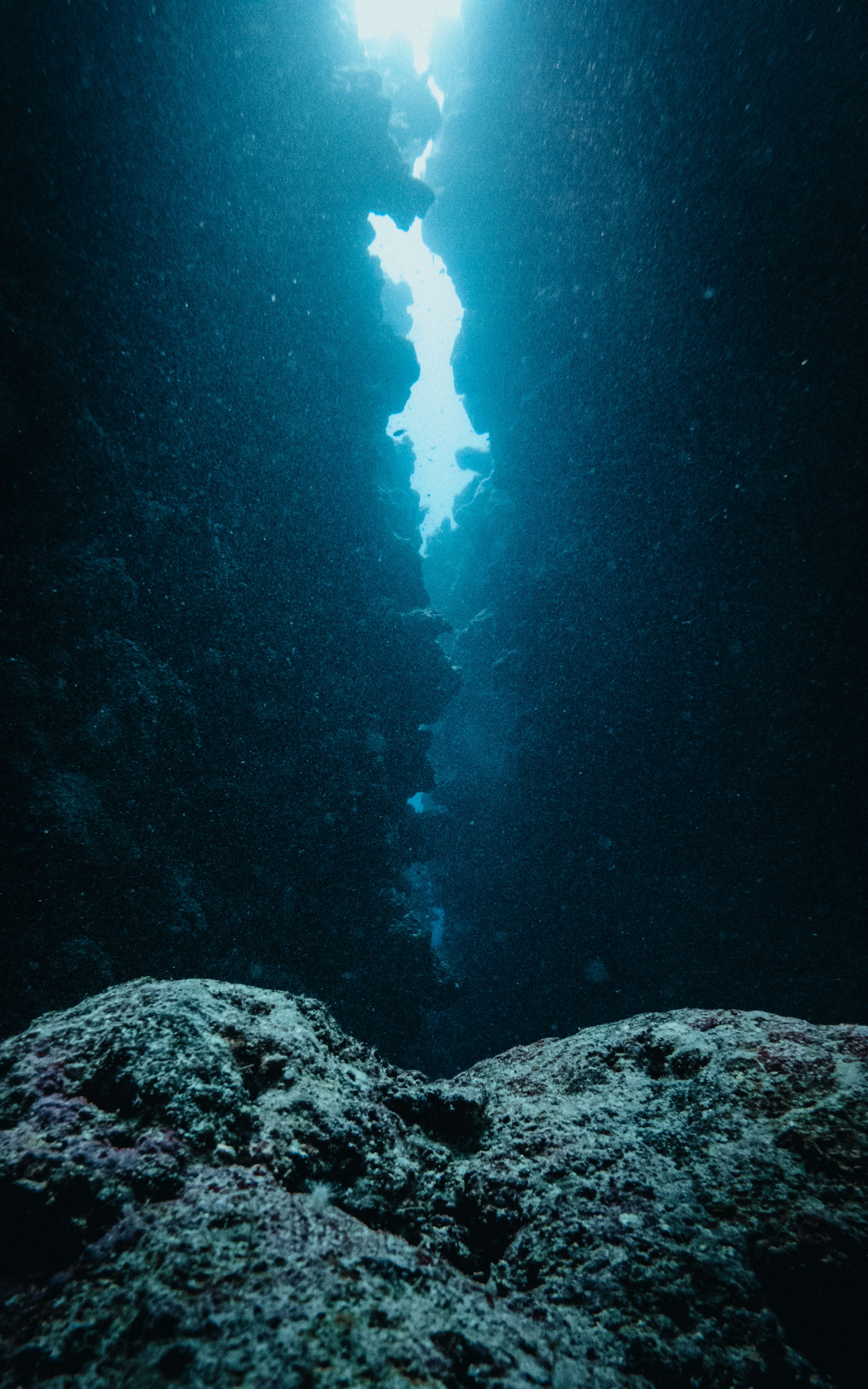 An underwater photograph looking straight up from the dark bottom of a narrow coral canyon. A sliver of bright blue surface water and intense sunlight breaks through the vertical split at the top, illuminating columns of floating sea particles.