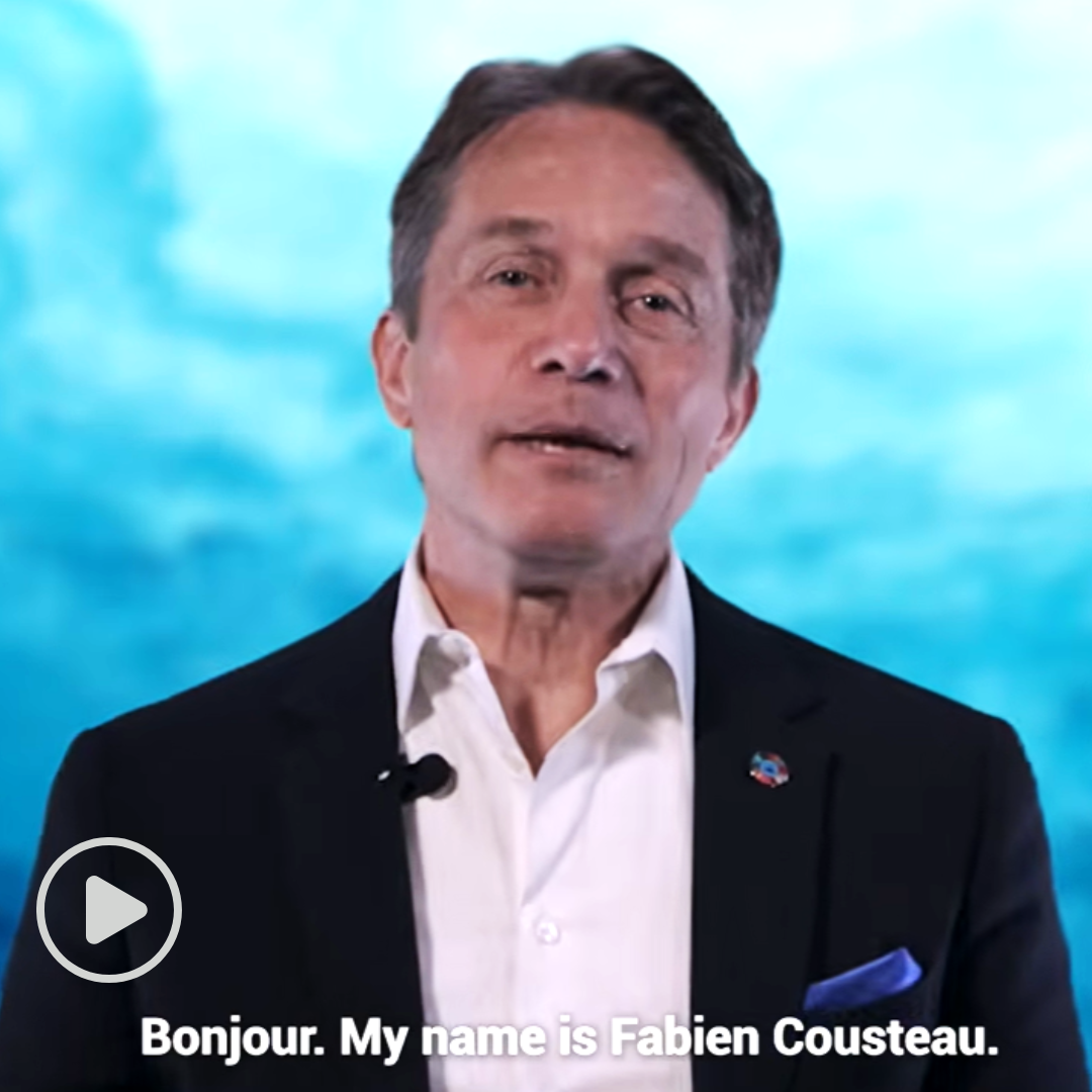 Fabien Cousteau in a dark suit jacket and white shirt speaking on camera against a blue ocean-themed background. White subtitle text at the bottom reads, 'Bonjour. My name is Fabien Cousteau."